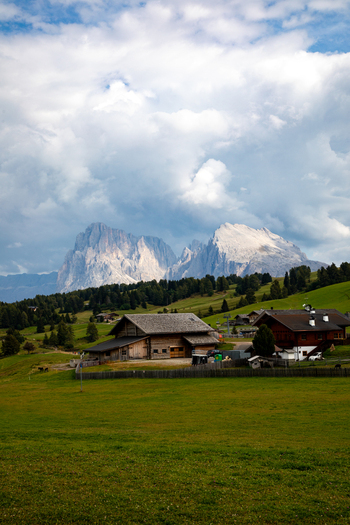 View of a traditional wooden alpine house in green meadows with the jagged peaks of the Dolomites under a cloudy sky in Rocca Pietore Veneto Italy. View of a traditional wooden alpine house in green meadows with the jagged peaks of the Dolomites under a cloudy sky in Rocca Pietore Veneto Italy.