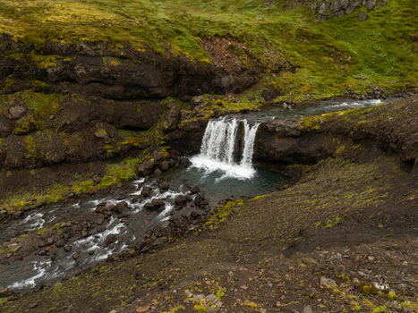 Aerial view of a waterfall cascading over dark volcanic rocks into a pool surrounded by vibrant green mossy terrain in the Eastern Region Iceland. Aerial view of a waterfall cascading over dark volcanic rocks into a pool surrounded by vibrant green mossy terrain in the Eastern Region Iceland.
