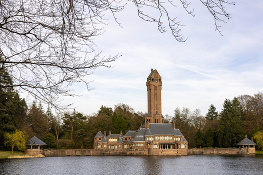 View of Country Residence Museum Jachthuis Sint Hubertus standing by a calm lake surrounded by dense forest under a clear sky Hoenderloo Gelderland Netherlands.
