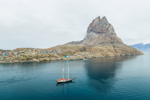 Aerial view of the iconic Uummannaq mountain rising above the colorful houses of the town and a sailing ship navigating the calm waters in Uummannaq Kitaa Greenland. Aerial view of the iconic Uummannaq mountain rising above the colorful houses of the town and a sailing ship navigating the calm waters in Uummannaq Kitaa Greenland.