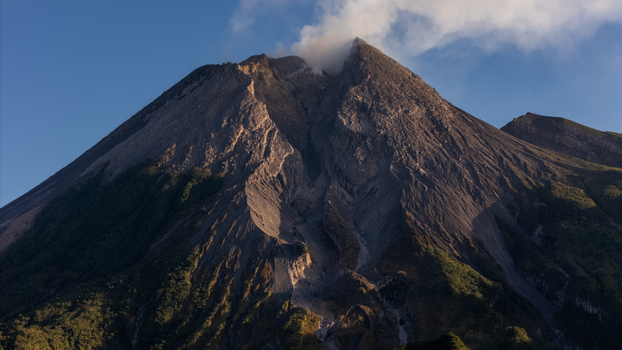 Aerial view of Mount Merapi with volcanic smoke rising from the crater rugged rocky slopes and lush green vegetation at the base under a clear blue sky in Indonesia.
