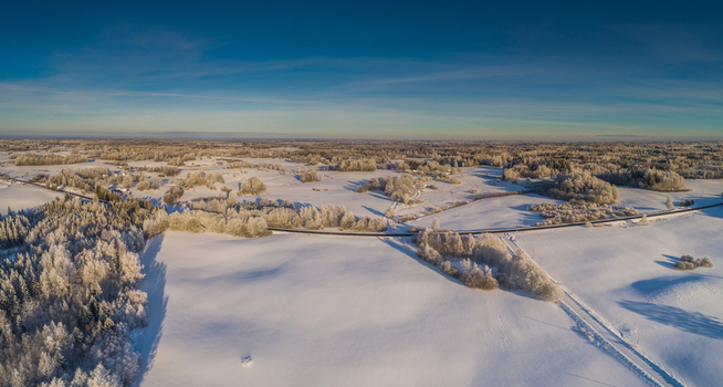 Aerial view of a vast snow covered landscape in Latvia featuring frost laden trees a winding road cutting through the winter fields and a clear blue sky. Aerial view of a vast snow covered landscape in Latvia featuring frost laden trees a winding road cutting through the winter fields and a clear blue sky.