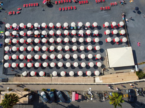 Aerial view of rows of white umbrellas and red sunbeds on a dark pebble beach with people walking and parked cars along the promenade in Imperia Liguria Italy. Aerial view of rows of white umbrellas and red sunbeds on a dark pebble beach with people walking and parked cars along the promenade in Imperia Liguria Italy.