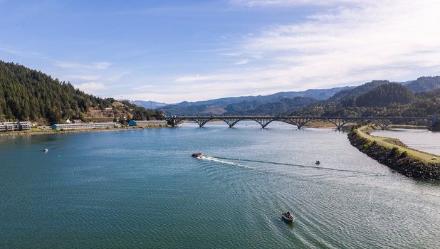 Aerial view of salmon fishing boats on the Rogue River near the Isaac Lee Patterson Bridge surrounded by forested hills under a clear blue sky in Gold Beach Oregon United States.