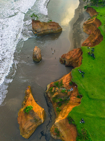 Aerial view of rugged coastal cliffs with green grassy slopes dark sand beach and crashing waves with people relaxing on the shore in Bali Bali Indonesia. Aerial view of rugged coastal cliffs with green grassy slopes dark sand beach and crashing waves with people relaxing on the shore in Bali Bali Indonesia.