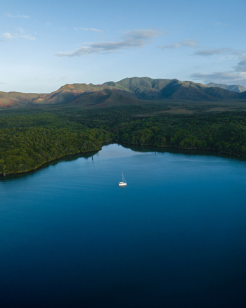 Aerial view of a solitary white sailboat glides through the deep blue waters fringed by vibrant green forest meeting rugged mountains Goro South Province New Caledonia. Aerial view of a solitary white sailboat glides through the deep blue waters fringed by vibrant green forest meeting rugged mountains Goro South Province New Caledonia.