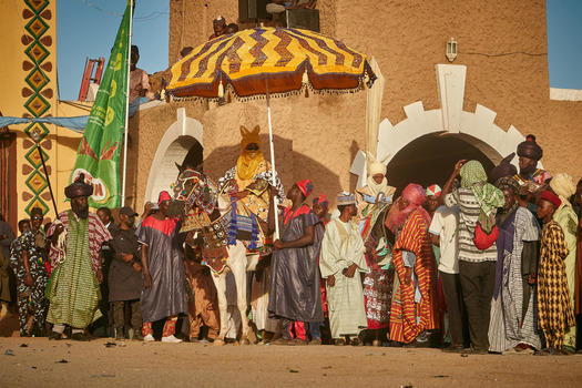 Zaria city Nigeria 22 March 2026 View of a vibrant procession with a regal figure on a horse beneath a striking umbrella passing an earthen toned building. Zaria city Nigeria 22 March 2026 View of a vibrant procession with a regal figure on a horse beneath a striking umbrella passing an earthen toned building.