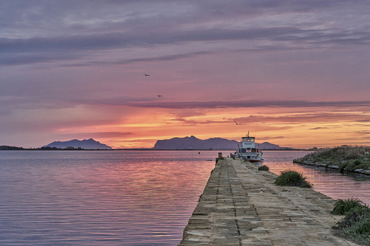 View of Saline dello Stagnone at sunset with a stone pier a small boat and distant mountains under a vibrant sky in Marsala Sicilia Italy.