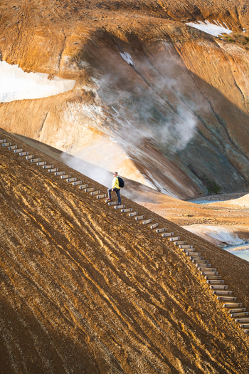 View of a hiker walking up a wooden staircase on the steep volcanic slopes of Kerlingarfjoll with geothermal steam and snow patches in Hrunamannahreppur Iceland.
