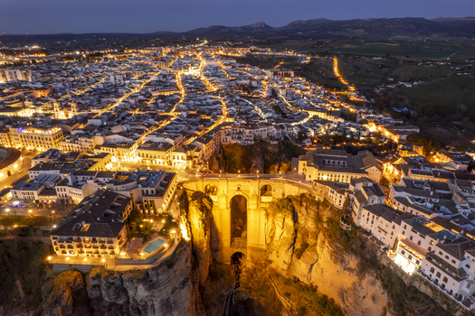 Aerial view of the illuminated Puente Nuevo bridge spanning the deep El Tajo gorge in Ronda with glowing streetlights illuminating the historic architecture. Ronda Andalusia Spain. Aerial view of the illuminated Puente Nuevo bridge spanning the deep El Tajo gorge in Ronda with glowing streetlights illuminating the historic architecture. Ronda Andalusia Spain.