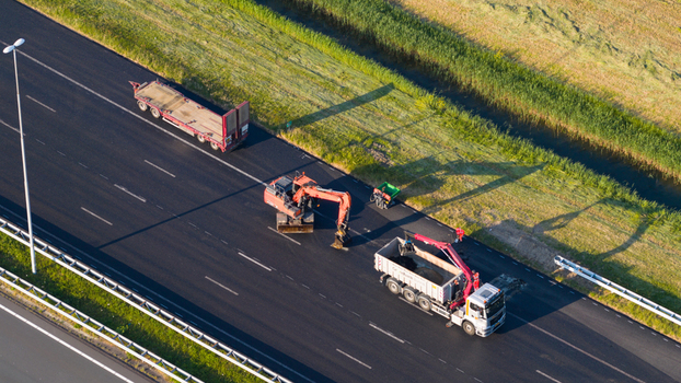 Waardenburg Netherlands 09 May 2026 Aerial view of road maintenance on a highway featuring an excavator and a truck on fresh asphalt next to green fields and a canal. Waardenburg Netherlands 09 May 2026 Aerial view of road maintenance on a highway featuring an excavator and a truck on fresh asphalt next to green fields and a canal.