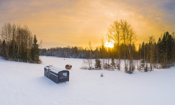 Aerial view of a modern cabin and a barrel sauna in a snow covered landscape with a fire pit under a golden sunset in Latvia. Aerial view of a modern cabin and a barrel sauna in a snow covered landscape with a fire pit under a golden sunset in Latvia.
