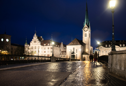 View of Fraumunster church illuminated at night with street lamps cobblestone bridge and pedestrians in Zurich Zurich Switzerland.