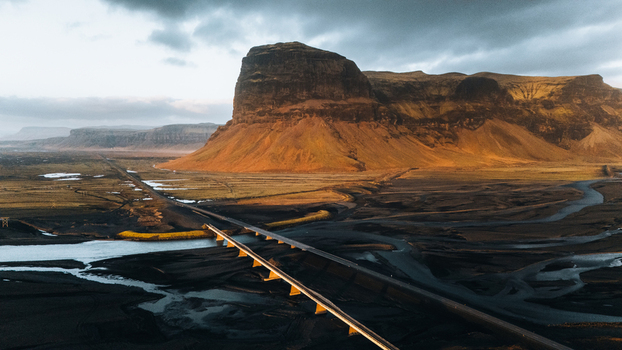 Aerial view of the massive Lomagnupur mountain and a long bridge crossing a glacial river delta under a dramatic cloudy sky in Skaftarhreppur Iceland.