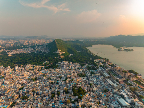 Aerial view of the historic city of Udaipur with dense urban rooftops Lake Pichola and surrounding green hills under the warm glow of the golden hour in Udaipur Rajasthan India. Aerial view of the historic city of Udaipur with dense urban rooftops Lake Pichola and surrounding green hills under the warm glow of the golden hour in Udaipur Rajasthan India.