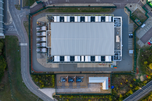 Aerial view of the Kao Data center facility featuring a large rectangular building with industrial cooling units and surrounding perimeter fencing in Harlow England United Kingdom. Aerial view of the Kao Data center facility featuring a large rectangular building with industrial cooling units and surrounding perimeter fencing in Harlow England United Kingdom.