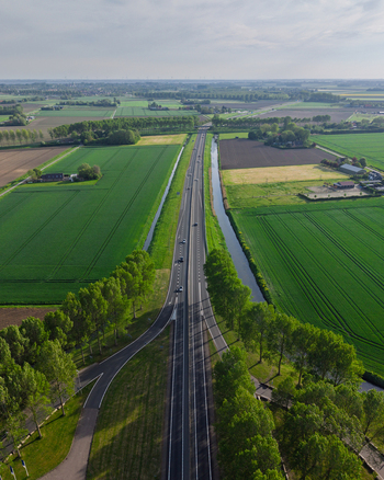 Aerial view of a straight highway with cars passing through vibrant green fields lined with tall trees and a parallel canal under a clear sky. Aerial view of a straight highway with cars passing through vibrant green fields lined with tall trees and a parallel canal under a clear sky.
