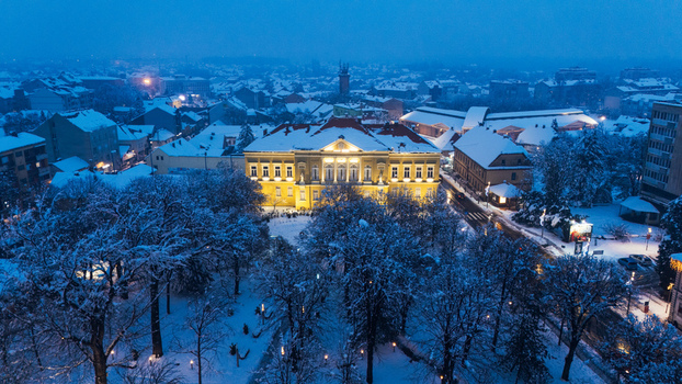Aerial view of the illuminated City Hall building surrounded by snow covered trees and rooftops under a twilight sky in Sremska Mitrovica Vojvodina Serbia.