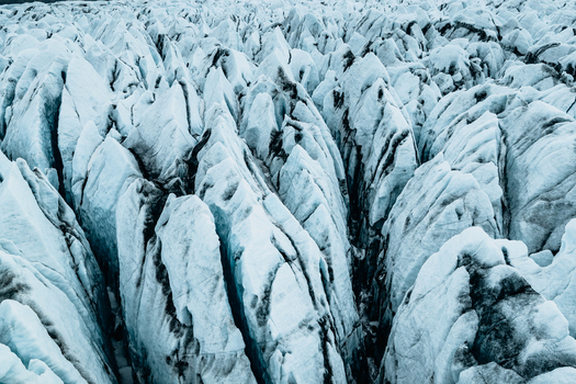 Aerial view of the rugged ice formations and deep crevasses of Vatnajokull glacier featuring intricate patterns of blue ice and dark volcanic ash deposits in Iceland.