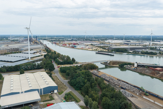 Aerial view of an industrial landscape in Genk featuring a canal with cargo barges wind turbines and warehouses surrounded by green trees in Genk Flanders Belgium. Aerial view of an industrial landscape in Genk featuring a canal with cargo barges wind turbines and warehouses surrounded by green trees in Genk Flanders Belgium.