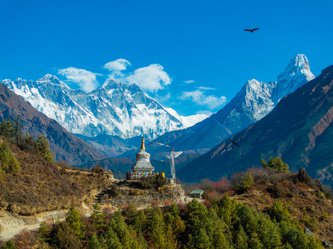 Aerial view of a Buddhist stupa perched on a hillside with snow capped Himalayan peaks in the background under a clear blue sky in Nepal.