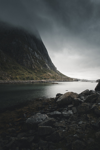 View of the dramatic Lofoten mountains rising above a calm fjord with a rugged rocky shoreline under a dark overcast sky in Nordland Norway. View of the dramatic Lofoten mountains rising above a calm fjord with a rugged rocky shoreline under a dark overcast sky in Nordland Norway.