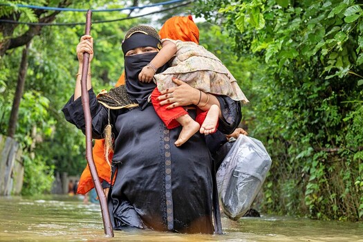 Banshkhali Upazila Bangladesh 10 July 2026 View of a woman in a black niqab wading through floodwaters while carrying a young child on her back and holding a wooden stick. Banshkhali Upazila Bangladesh 10 July 2026 View of a woman in a black niqab wading through floodwaters while carrying a young child on her back and holding a wooden stick.