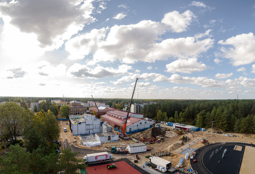 Aerial view of a construction site featuring a tall crane partially built structures and heavy machinery surrounded by a dense forest under a cloudy blue sky in Latvia. Aerial view of a construction site featuring a tall crane partially built structures and heavy machinery surrounded by a dense forest under a cloudy blue sky in Latvia.