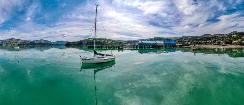 Aerial view of a sailboat anchored in the calm turquoise waters of Akaroa harbor with a wooden pier and buildings along the coastline in Akaroa Canterbury Region New Zealand.