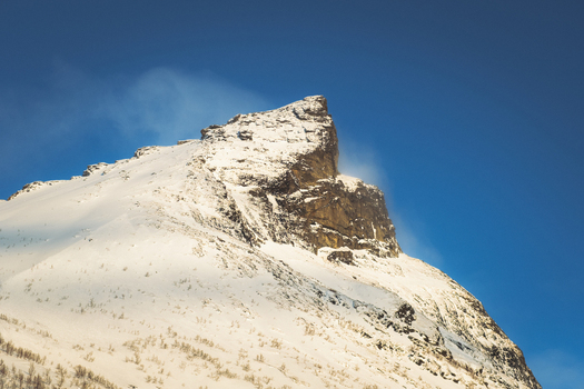 View of a snow dusted mountain peak rises majestically against a clear vibrant blue sky a contrast of cold whites and warm stone textures Indregård Troms Norway. View of a snow dusted mountain peak rises majestically against a clear vibrant blue sky a contrast of cold whites and warm stone textures Indregård Troms Norway.