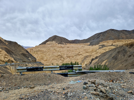 View of the Penzi La road winding through the rugged arid mountains of the Zanskar valley with a metal guardrail in the foreground under a cloudy sky in Zanskar Ladakh India.