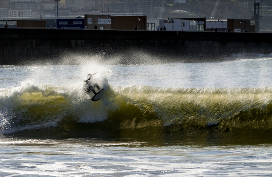 View of a surfer riding a breaking wave at Ereaga Beach with water spray glistening in the sunlight and an urban backdrop in Algorta Euskadi Spain.
