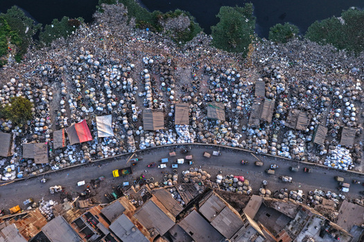 Aerial view of the Buriganga River bank featuring a dense collection of white sacks small makeshift structures and a busy road with vehicles in Dhaka Dhaka Division Bangladesh. Aerial view of the Buriganga River bank featuring a dense collection of white sacks small makeshift structures and a busy road with vehicles in Dhaka Dhaka Division Bangladesh.