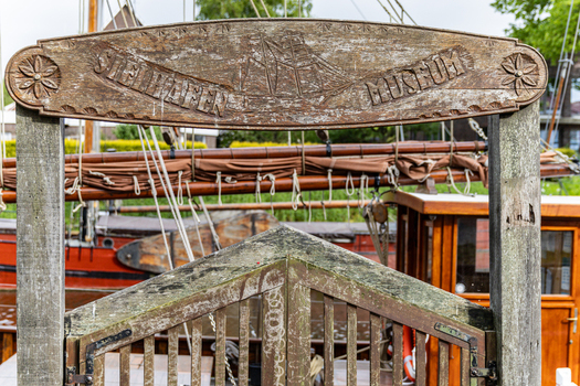 View of the wooden entrance sign of the Sielhafen Museum with a traditional sailing boat in the background at Hafen Harlesiel in Wittmund Lower Saxony Germany. View of the wooden entrance sign of the Sielhafen Museum with a traditional sailing boat in the background at Hafen Harlesiel in Wittmund Lower Saxony Germany.