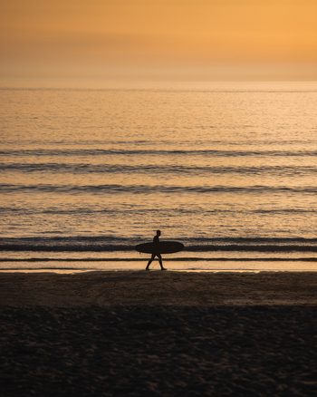 Penzance England United Kingdom 18 July 2021 View of a surfer in silhouette carrying a surfboard along the sandy shore of Gwenver Beach during a golden sunset.