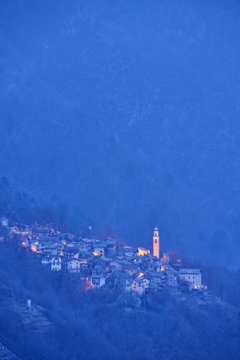 View of the historic village of Valchiavenna illuminated at dusk with stone houses a bell tower and warm lights glowing against the blue twilight in Lombardia Italy.