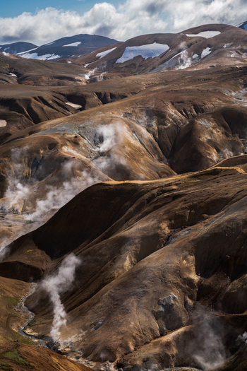 View of geothermal steam rising from rugged brown hillsides a stark contrast to the distant snow capped mountains under a bright blue sky Hrunamannahreppur Iceland.