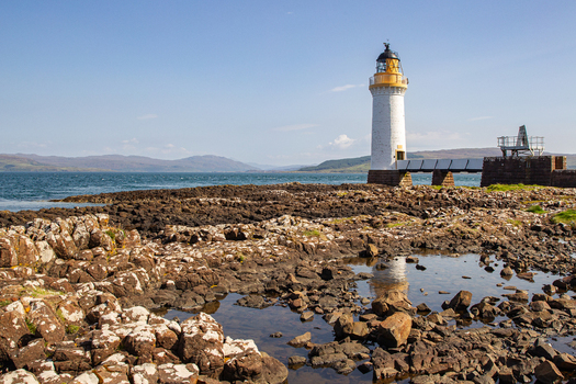 View of the Tobermory Lighthouse standing on a rocky shoreline with distant hills and a clear blue sky Isle of Mull Scotland United Kingdom. View of the Tobermory Lighthouse standing on a rocky shoreline with distant hills and a clear blue sky Isle of Mull Scotland United Kingdom.