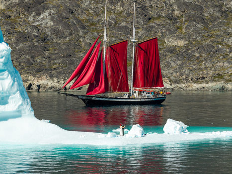 Aerial view of a sailboat with striking red sails navigating icy waters near a large iceberg with a couple standing on the ice in Greenland. Aerial view of a sailboat with striking red sails navigating icy waters near a large iceberg with a couple standing on the ice in Greenland.