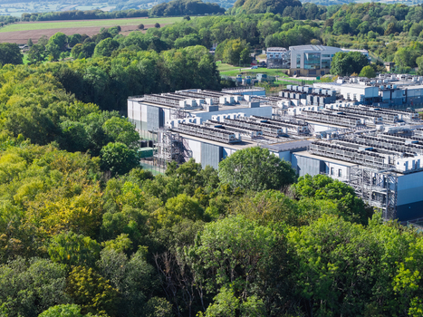Aerial view of a modern data center facility nestled within a dense forest featuring industrial cooling units on the roof and surrounding green trees in the United Kingdom.