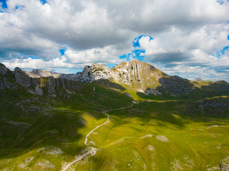 Aerial view of Bobotov Kuk featuring rugged mountain peaks lush green slopes and a winding trail under a cloudy blue sky in Zabljak Municipality Montenegro. Aerial view of Bobotov Kuk featuring rugged mountain peaks lush green slopes and a winding trail under a cloudy blue sky in Zabljak Municipality Montenegro.