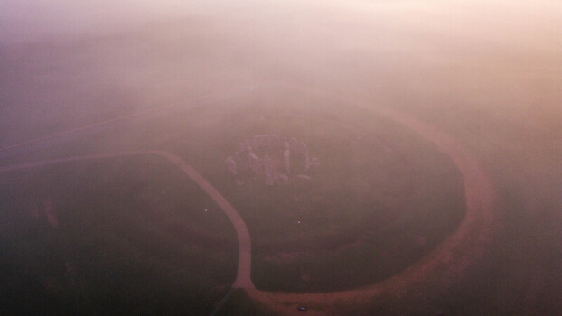 Aerial view of the ancient stone circle of Stonehenge surrounded by thick morning mist and soft golden light in the grassy landscape of Salisbury England United Kingdom. Aerial view of the ancient stone circle of Stonehenge surrounded by thick morning mist and soft golden light in the grassy landscape of Salisbury England United Kingdom.