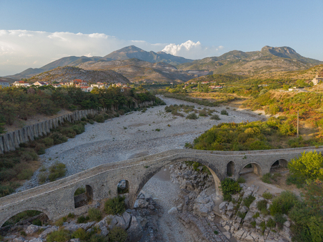 Aerial view of the historic Mesi Bridge spanning a dry riverbed with stone arches surrounded by rolling hills and lush vegetation under a clear sky in Boks Qarku i Shkodres Albania.