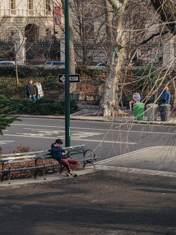 New York New York United States 13 April 2015 View of a young man sitting on a park bench using his smartphone with people walking along a street and bare trees in the background.