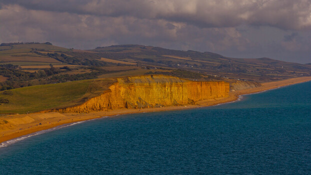 Aerial view of the iconic golden sandstone cliffs of the Jurassic Coast meeting the blue sea under a dramatic cloudy sky in West Bay England United Kingdom. Aerial view of the iconic golden sandstone cliffs of the Jurassic Coast meeting the blue sea under a dramatic cloudy sky in West Bay England United Kingdom.