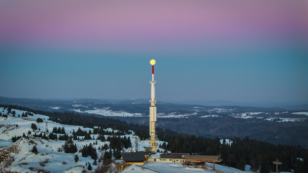 Aerial view of a tall white and red telecommunications tower piercing the pastel dawn sky over a snow covered landscape Gex Auvergne Rh&ocirc;ne Alpes France.
