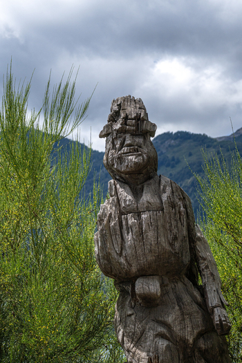 View of a weathered wooden sculpture of a person standing amidst green bushes with mountains in the background at Bahia Brava Villa La Angostura Neuquen Province Argentina. View of a weathered wooden sculpture of a person standing amidst green bushes with mountains in the background at Bahia Brava Villa La Angostura Neuquen Province Argentina.