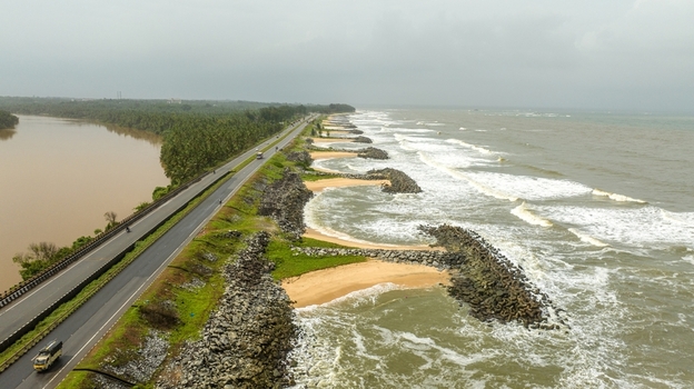 Aerial view of Maravanthe beach with a road running between the river and the Arabian Sea featuring rock breakwaters and lush green vegetation in Maravanthe Karnataka India. Aerial view of Maravanthe beach with a road running between the river and the Arabian Sea featuring rock breakwaters and lush green vegetation in Maravanthe Karnataka India.