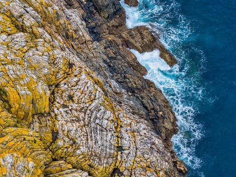 Aerial view of Silence Beach Viewpoint featuring rugged textured cliffs covered in yellow lichen and deep blue ocean waves crashing against the rocks in Cudillero Asturias Spain.