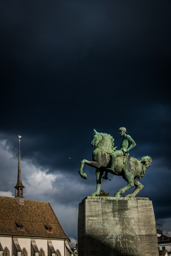 Zurich Switzerland 02 April 2023 View of a bronze equestrian statue rises against a dramatic dark sky juxtaposed with the warm tones of a nearby church roof. Zurich Switzerland 02 April 2023 View of a bronze equestrian statue rises against a dramatic dark sky juxtaposed with the warm tones of a nearby church roof.